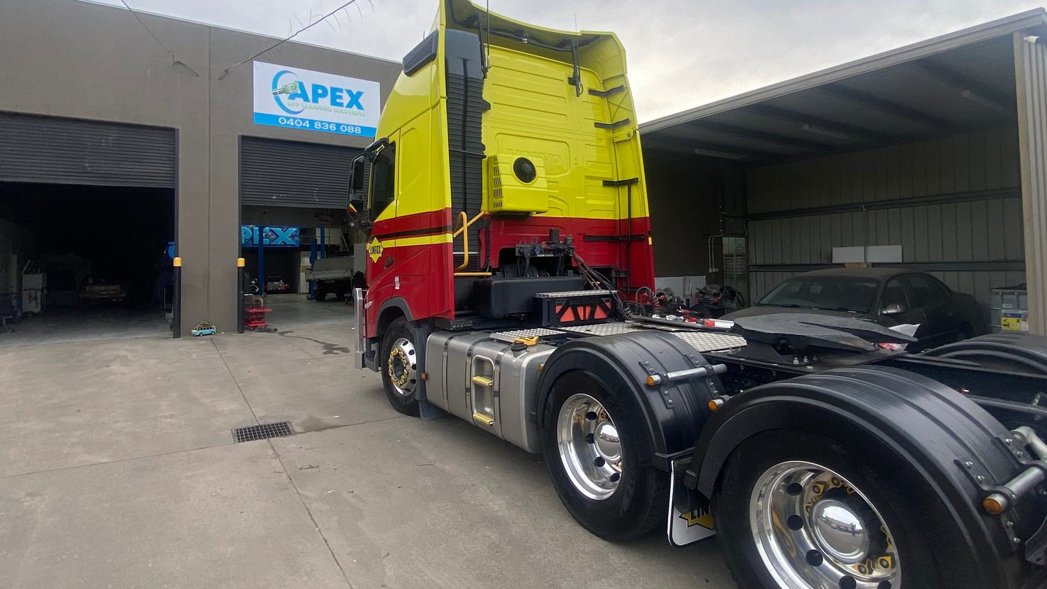 A yellow and red prime mover parked outside the Apex workshop, with the Apex DPF Cleaning Solutions sign above the roller doors.